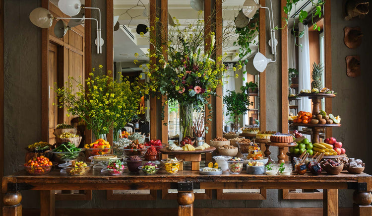 Alavya Hotel's abundant breakfast buffet table, photographed for the Montgomery Press hotel book. A long wooden table is laden with fresh fruit, vegetables, pastries, nuts and towering flower arrangements — vibrant food photography capturing the farm-to-table philosophy of this award-winning boutique hotel in Alacati, Turkey.