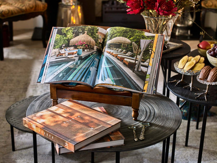 The Alavya hotel book, produced by Montgomery Press, displayed open on a wooden book stand in the hotel's sitting room. The pages show the iconic pool photography. Closed copies of the hardback book with its warm timber-effect cover sit beneath, alongside madeleines, fresh fruit and a magnifying glass. A bespoke luxury coffee table book commission for an award-winning boutique hotel on the Aegean coast of Turkey.