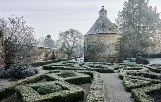 A frosty garden scene with trimmed hedges, a gravel path, bare trees, evergreen trees, and a round stone building with a conical roof under a pale sky. Photograph by Andrew Montgomery.