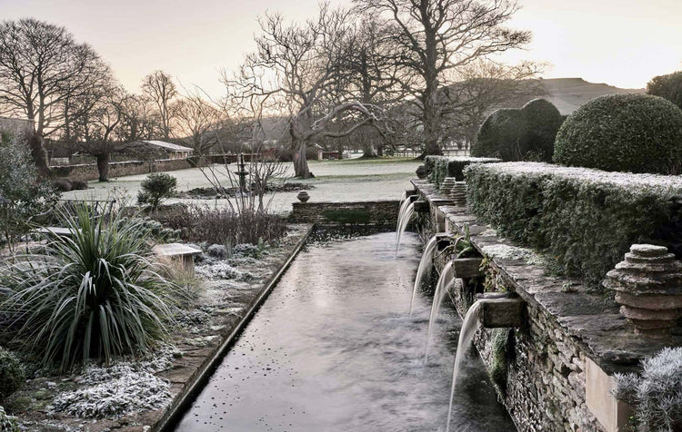 A frosty stone garden scene with a long narrow pool, small cascading water features, trimmed hedges, leafless trees, and a pale sky at sunrise or sunset. Photograph by Andrew Montgomery.